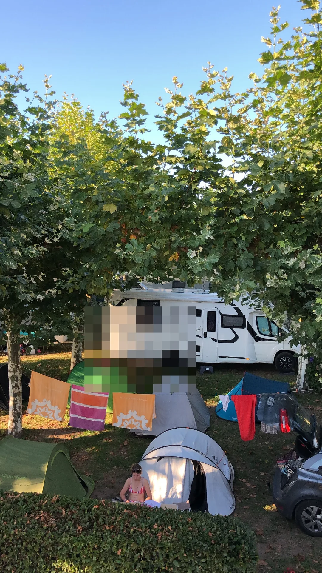 Zona de camping con autocaravana, tiendas y ropa tendida entre árboles.