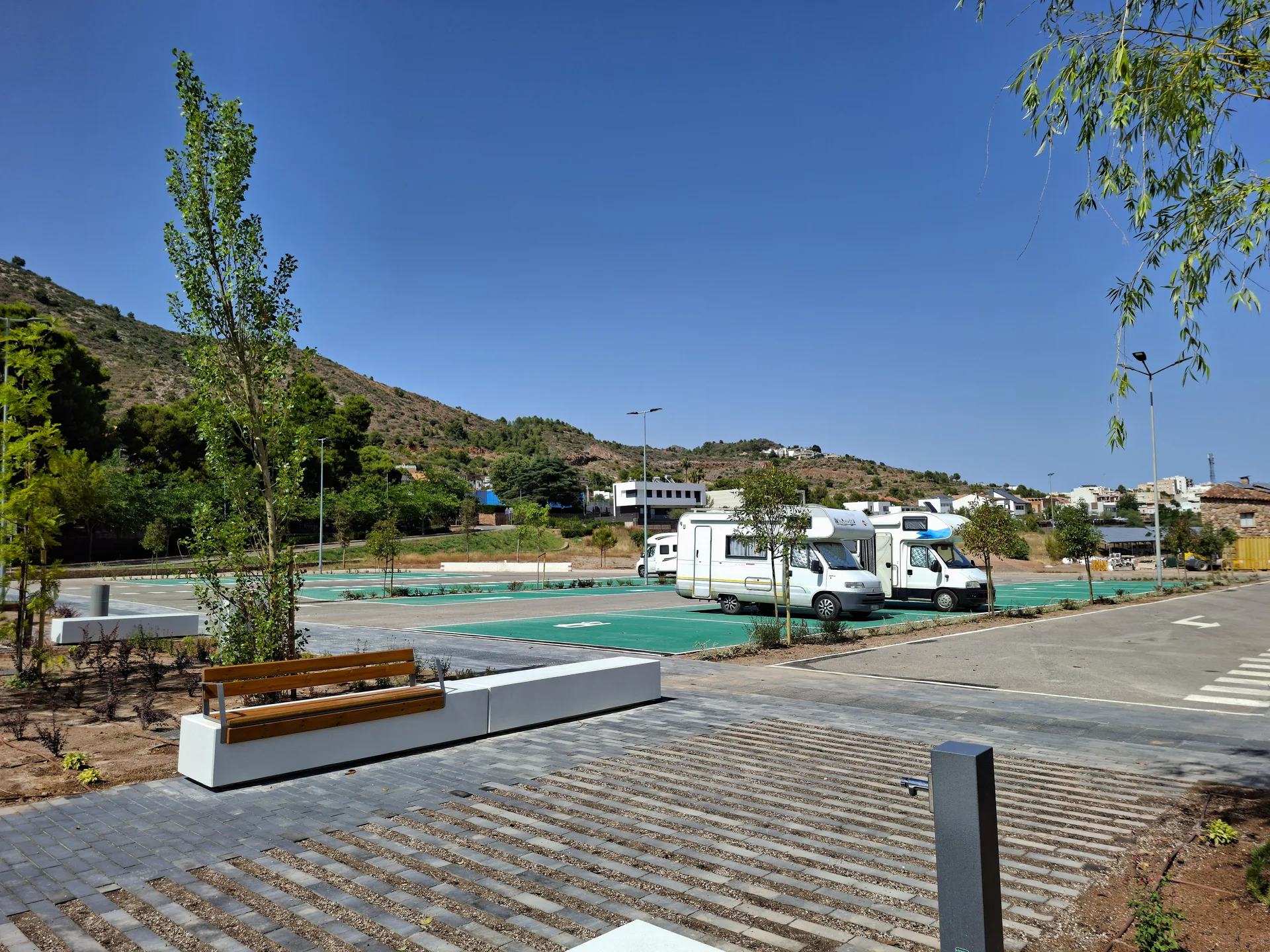 Área de autocaravanas con plazas pintadas, bancos y varias campers estacionadas, en un entorno abierto con colinas.