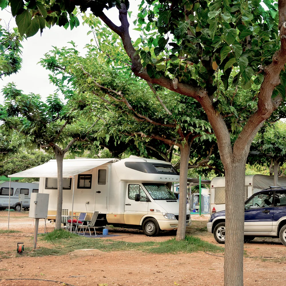 Área de camping con autocaravana bajo árboles y varias parcelas ocupadas.