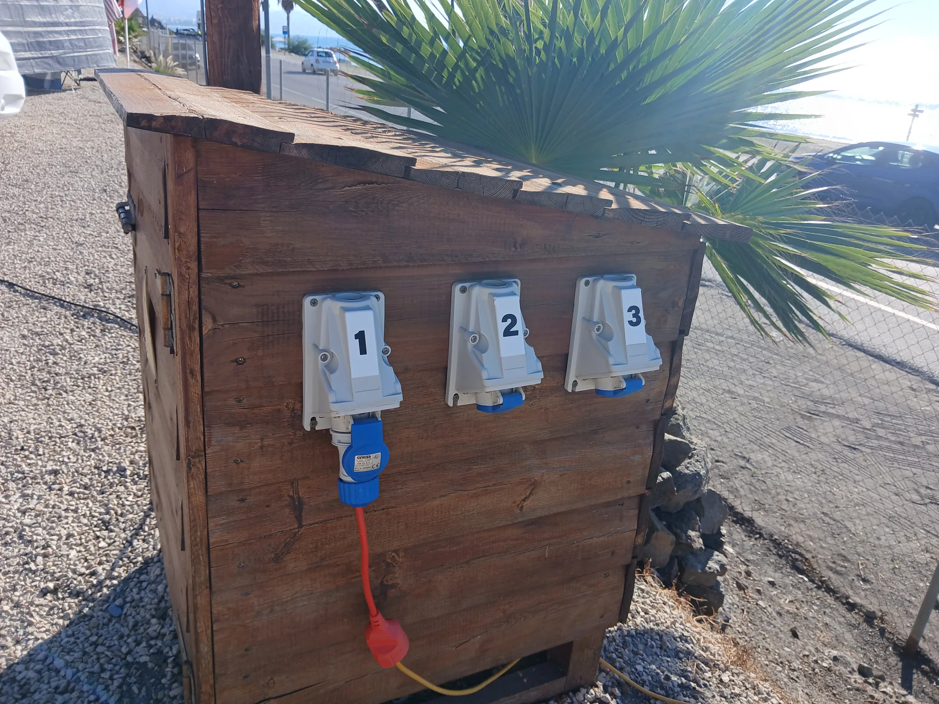 Punto de toma eléctrica exterior numerado junto al área, con vistas al paseo y la costa.