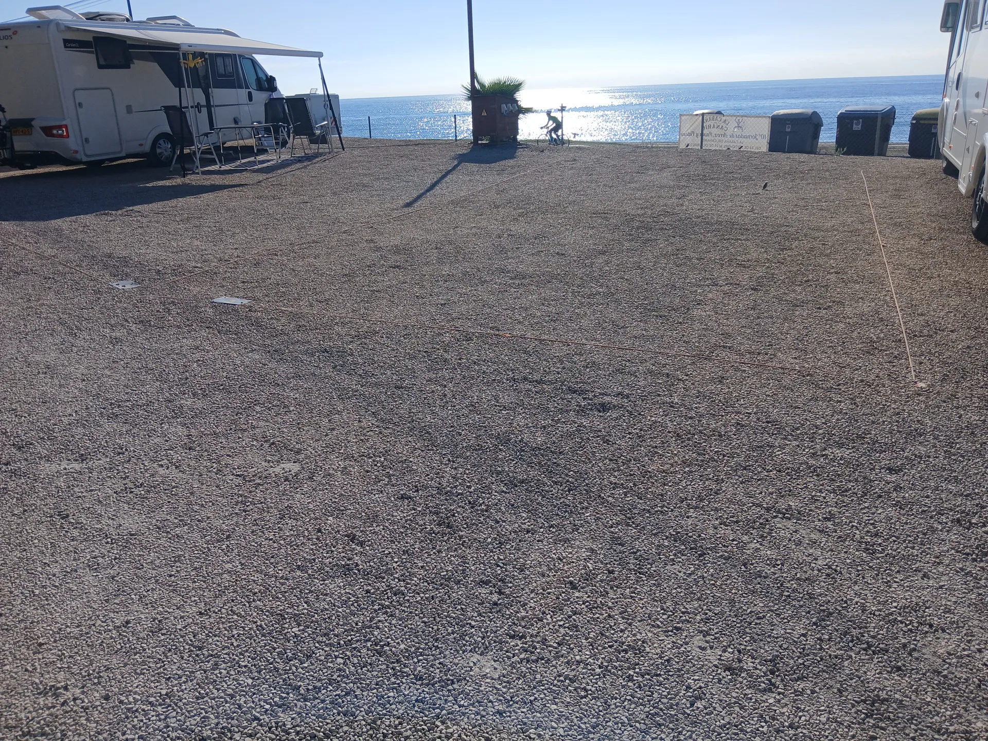 Área de autocaravanas junto al mar con parcelas de grava y vehículos estacionados.