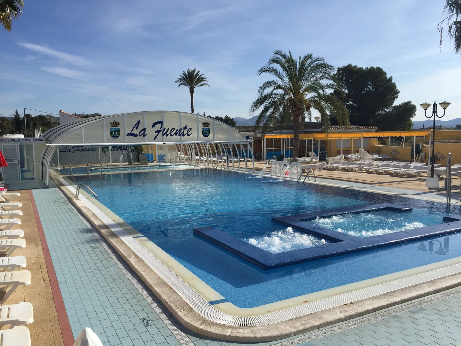 Piscina termal exterior de Camping Fuente en Los Baños de Fortuna, con la cubierta abovedada, hamacas y palmeras