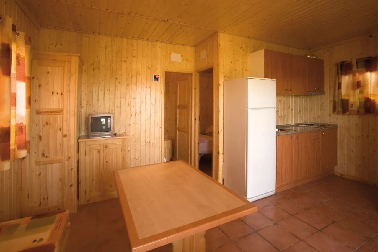 Interior de una cabaña/estancia del camping con cocina, mesa, nevera y armarios.